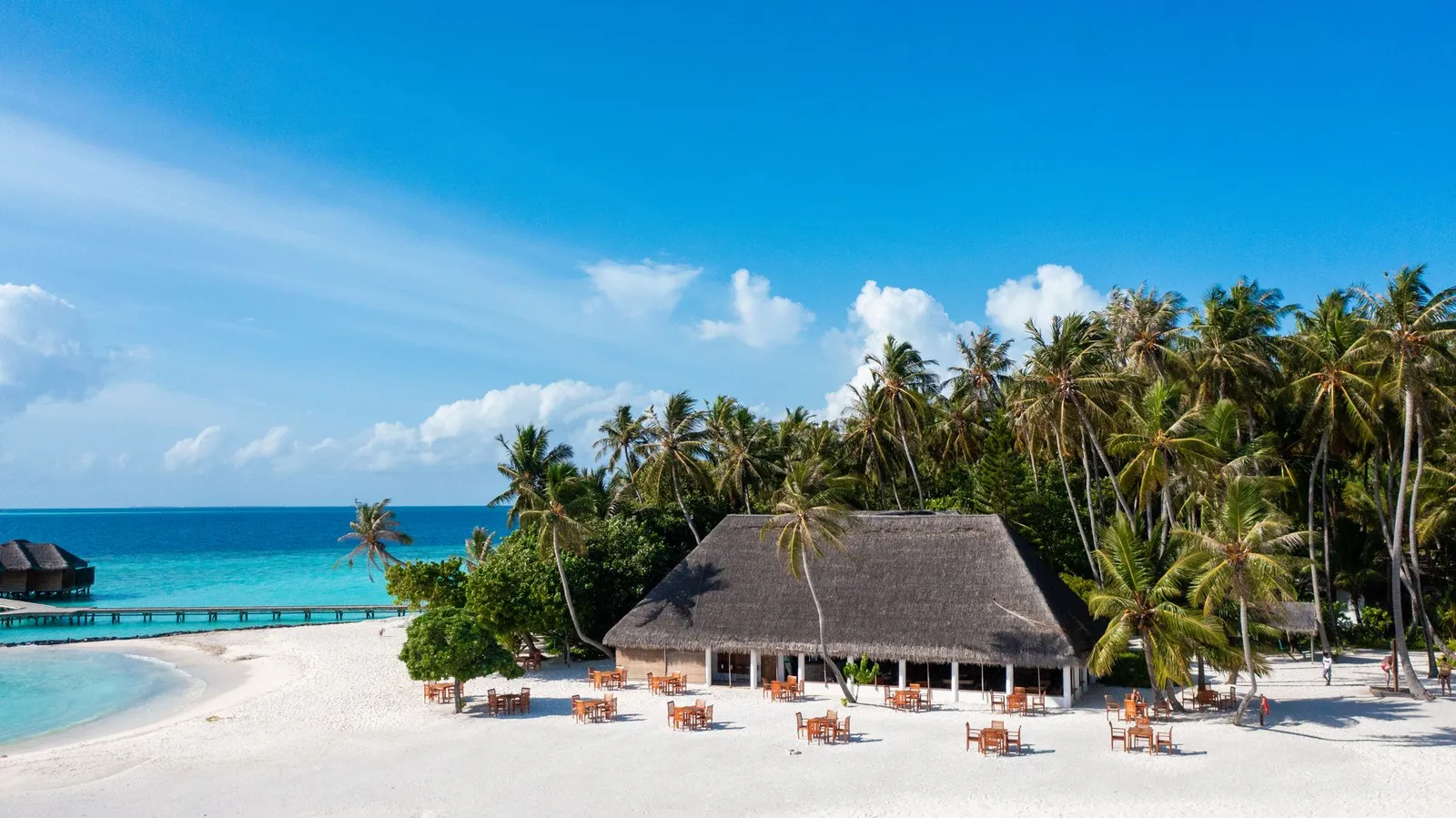 A thatched building on the beach, its tables on the sand, tall palms and the water bungalows’ jetty on the left.