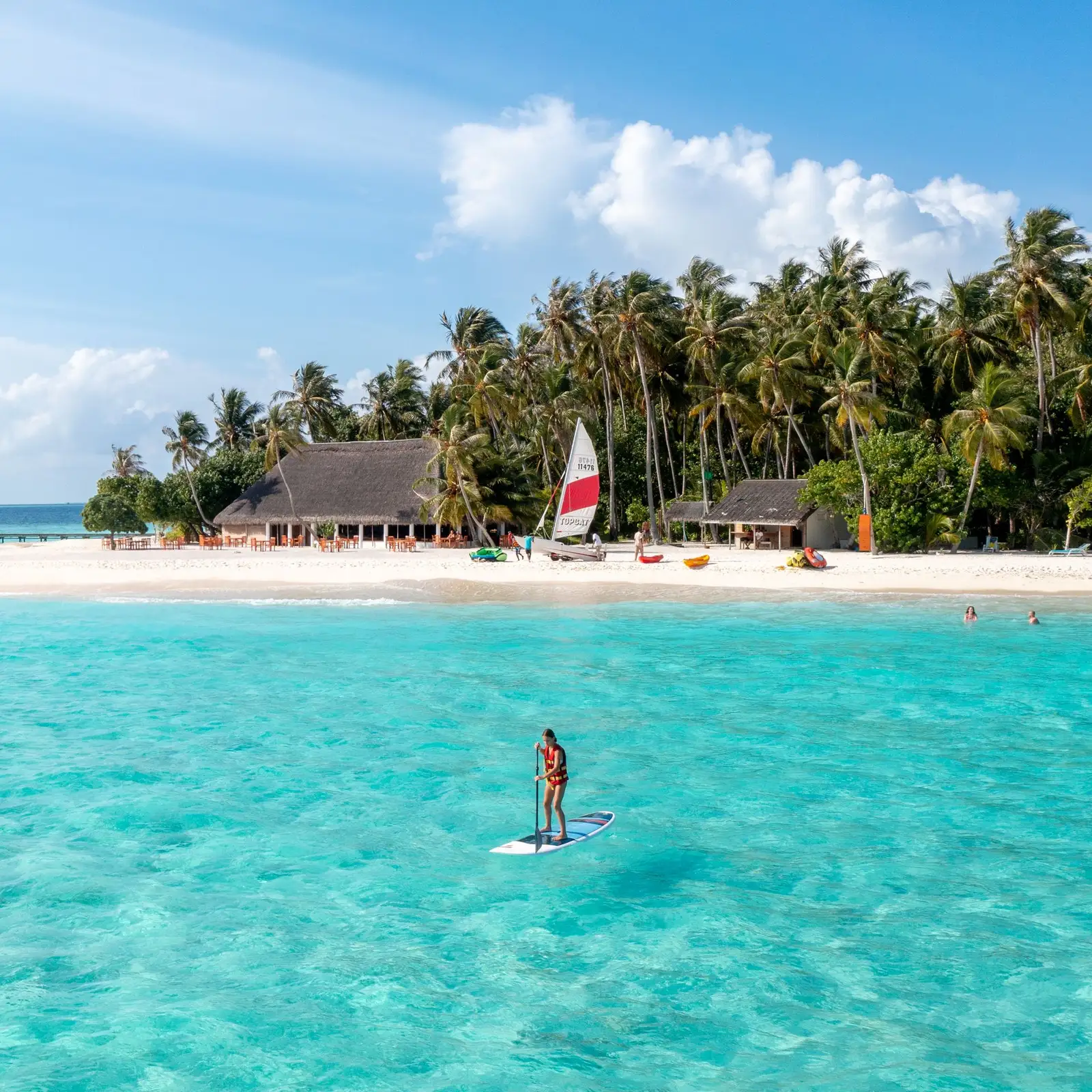 Someone standing on a paddleboard in the lagoon, with the beach, a catamaran and the palms behind.