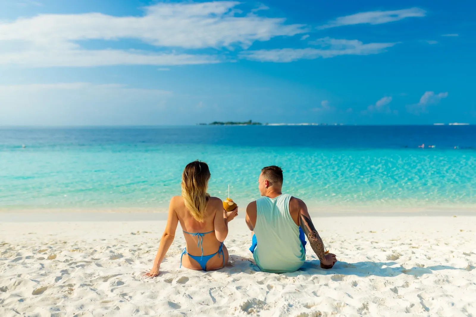 A couple sitting on the white sand holding 2 coconuts, facing the turquoise sea and a distant island.