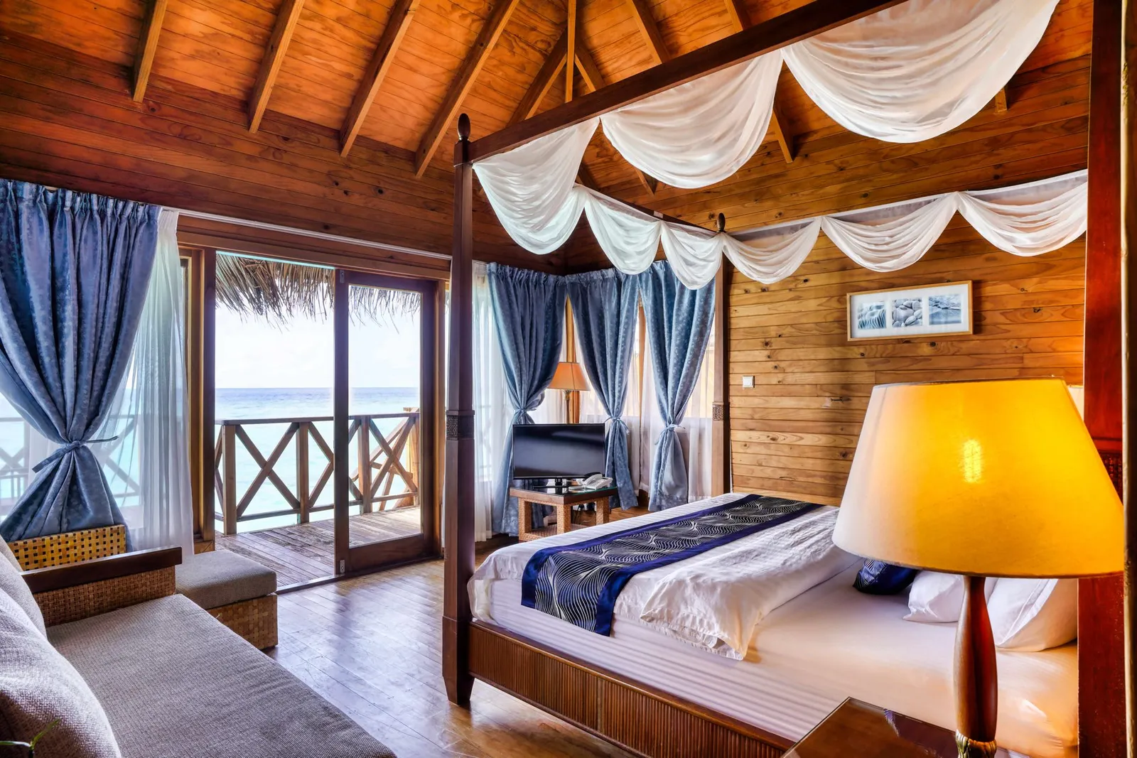 A Water Villa bedroom: the four-poster bed with white drapes, the timber walls and the door open onto the deck.