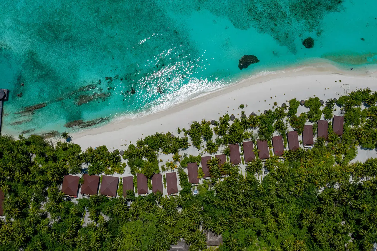 Filitheyo’s beach from above: the row of villa roofs among the trees, the white sand and the reef beneath the lagoon.