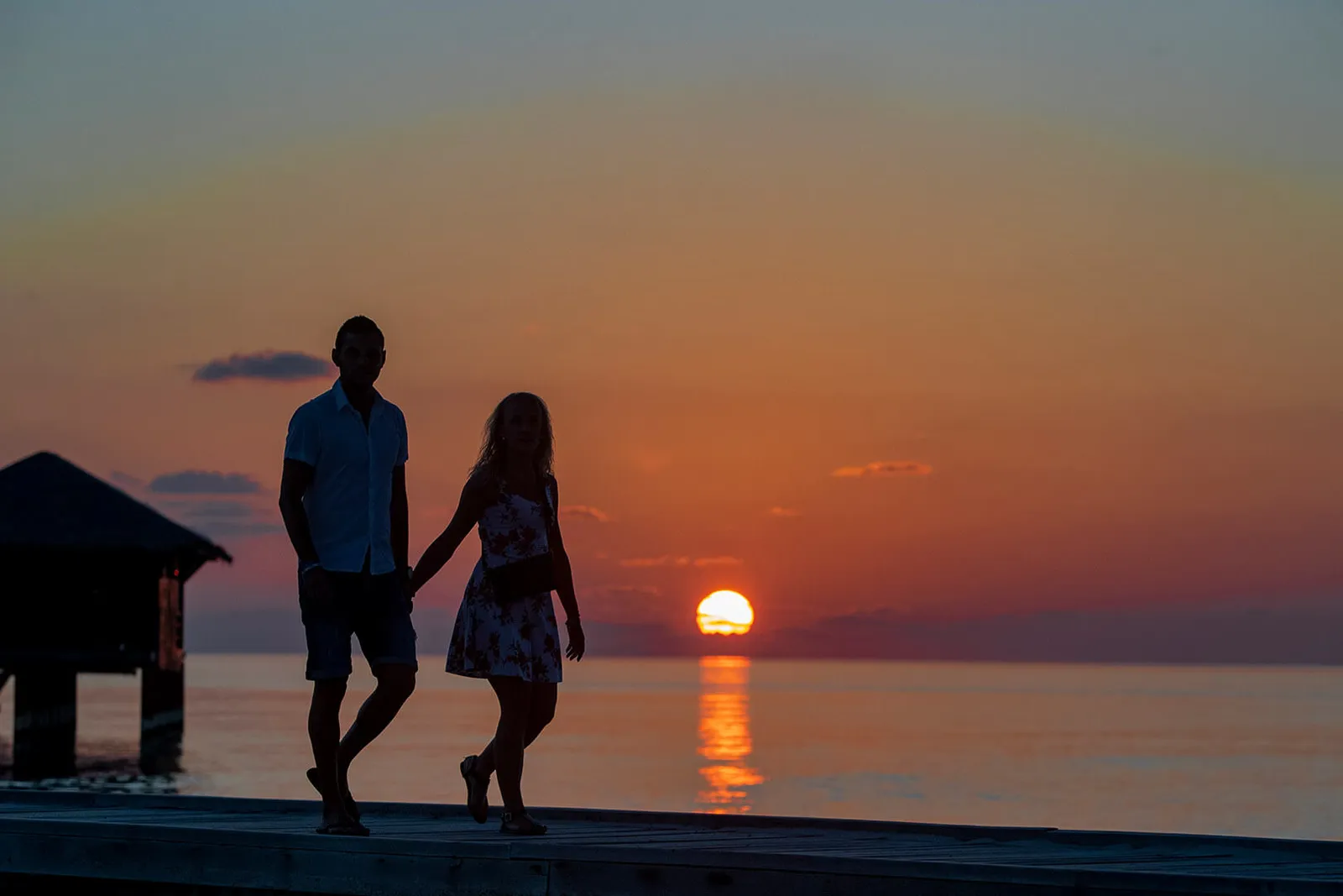 A couple walk hand in hand along the water villa jetty, in silhouette, as the sun meets the sea.