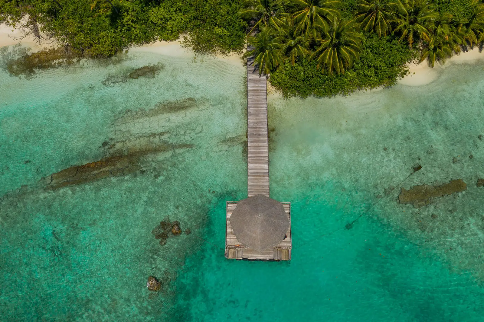 The wooden jetty running from the beach into the lagoon, seen from above, with the dark patches of reef in the clear water.