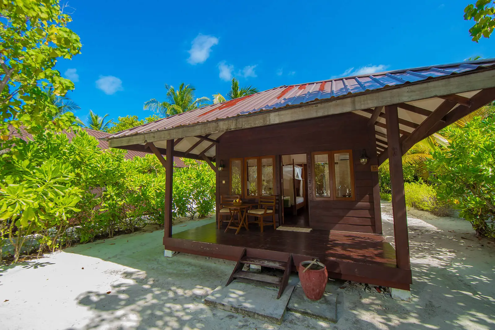 A detached Deluxe Villa among the shrubs, in dark timber with a red roof, its veranda and the sand in front.