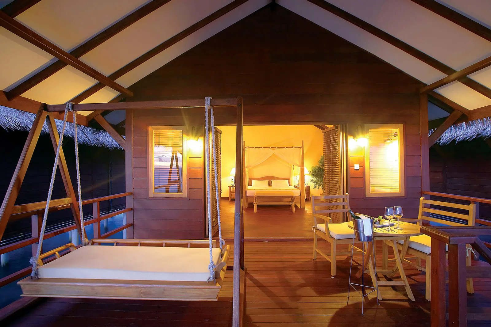 A Water Villa’s covered deck at dusk, with the udoli swing seat and the four-poster bed lit up inside.