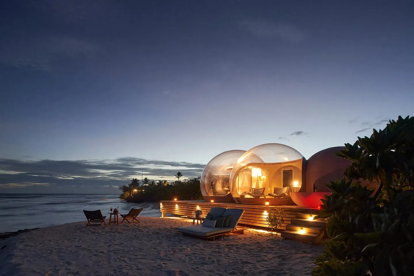 The Beach Bubble lit up on the sand at dusk, the sea behind