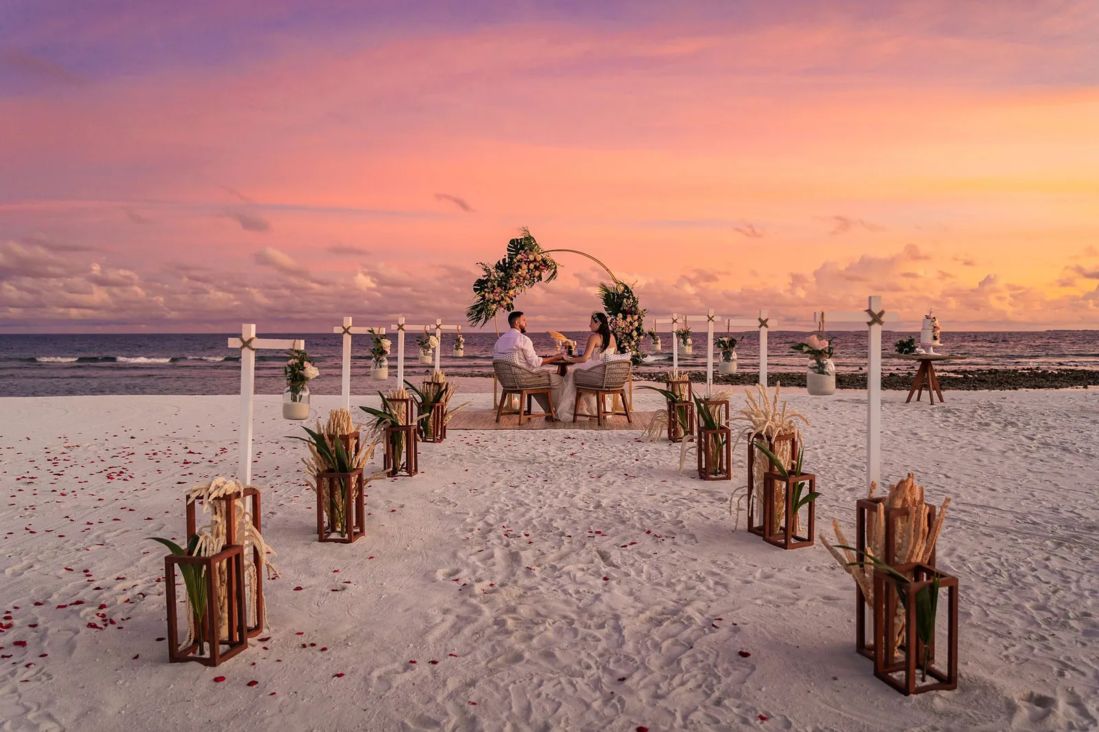 A couple dining on the beach at sunset, at the end of an aisle of lanterns beneath a floral arch