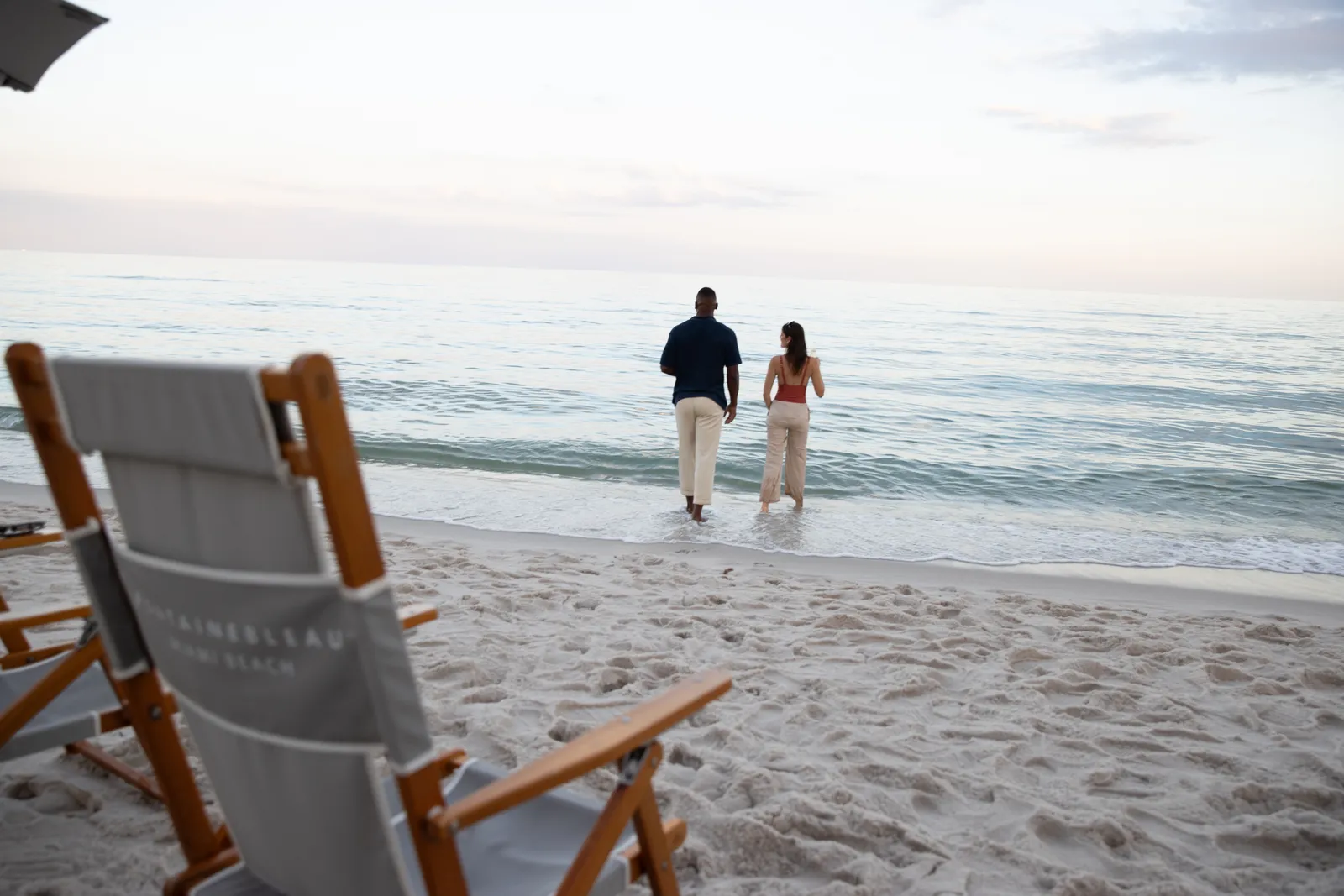 A wood and canvas chair on the sand with the hotel’s name on its back and, further off, a couple standing in the shallows.