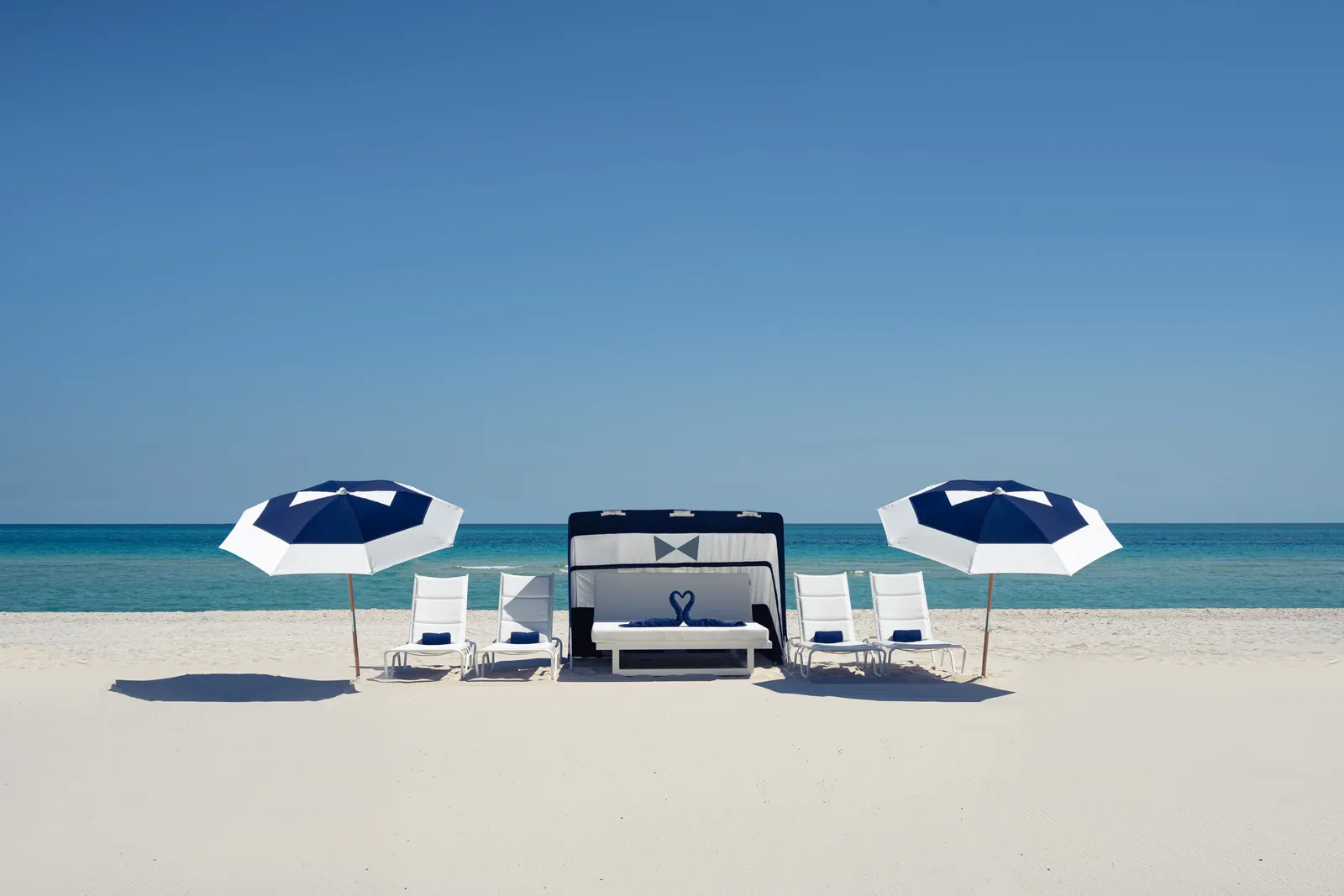 The beach in front of the hotel: white loungers, 2 blue-and-white umbrellas and a beach bed on pale sand, with the turquoise Atlantic behind.