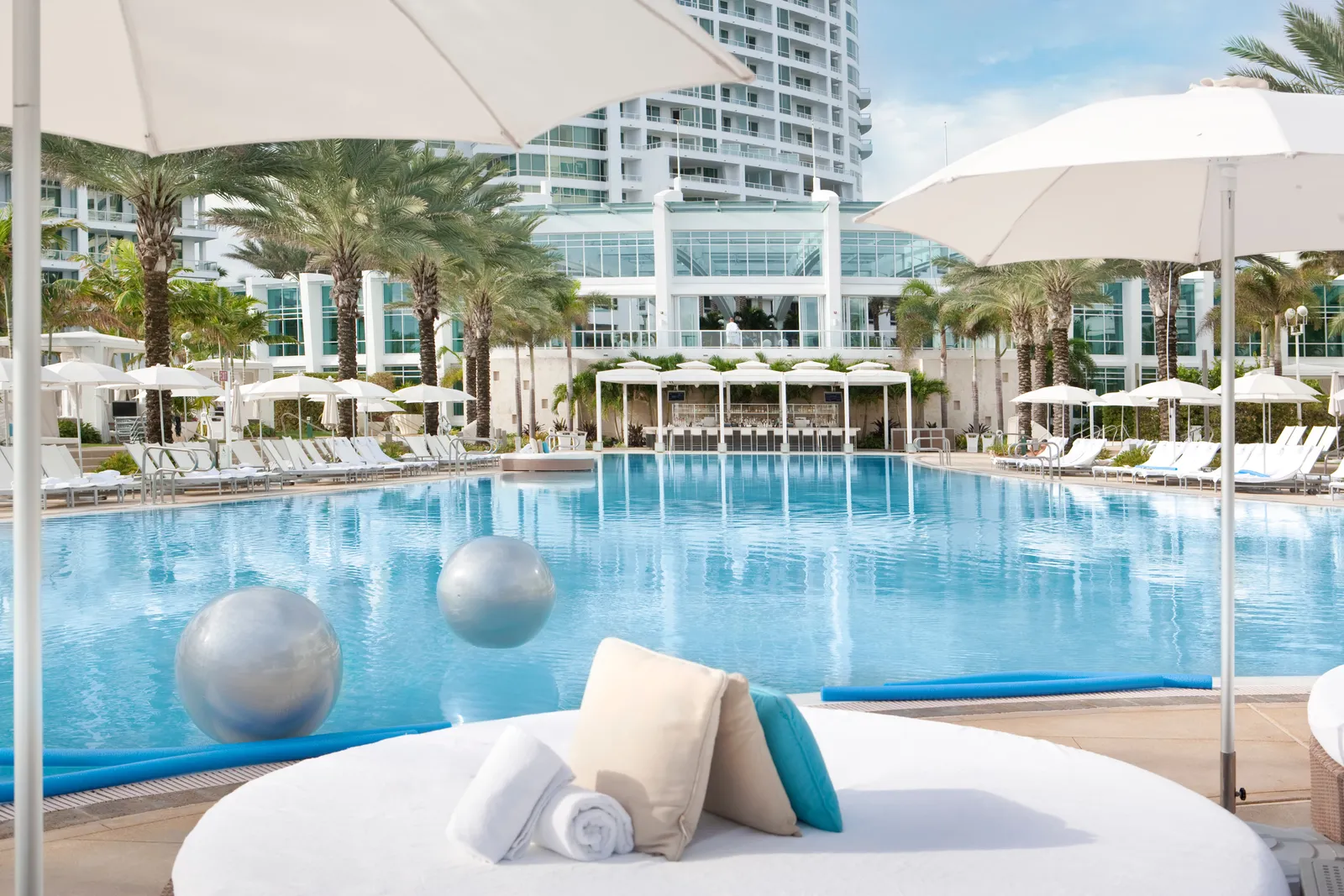 A round daybed with towels and cushions at the edge of the main pool, among white umbrellas, palms and the hotel’s towers.