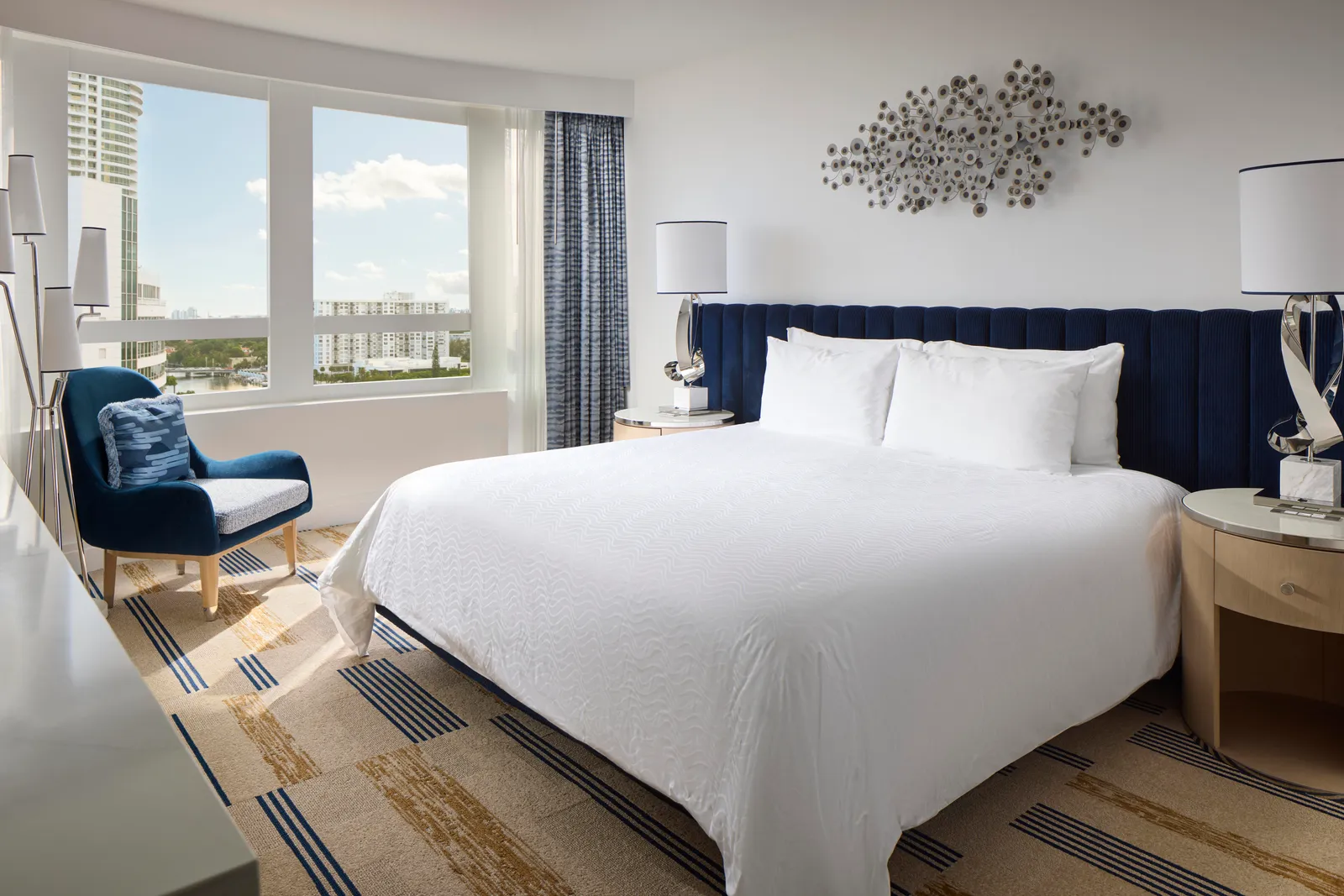 A Bay View Junior Suite with Balcony: the king bed with its blue headboard, an armchair and the windows onto Miami Beach’s towers.