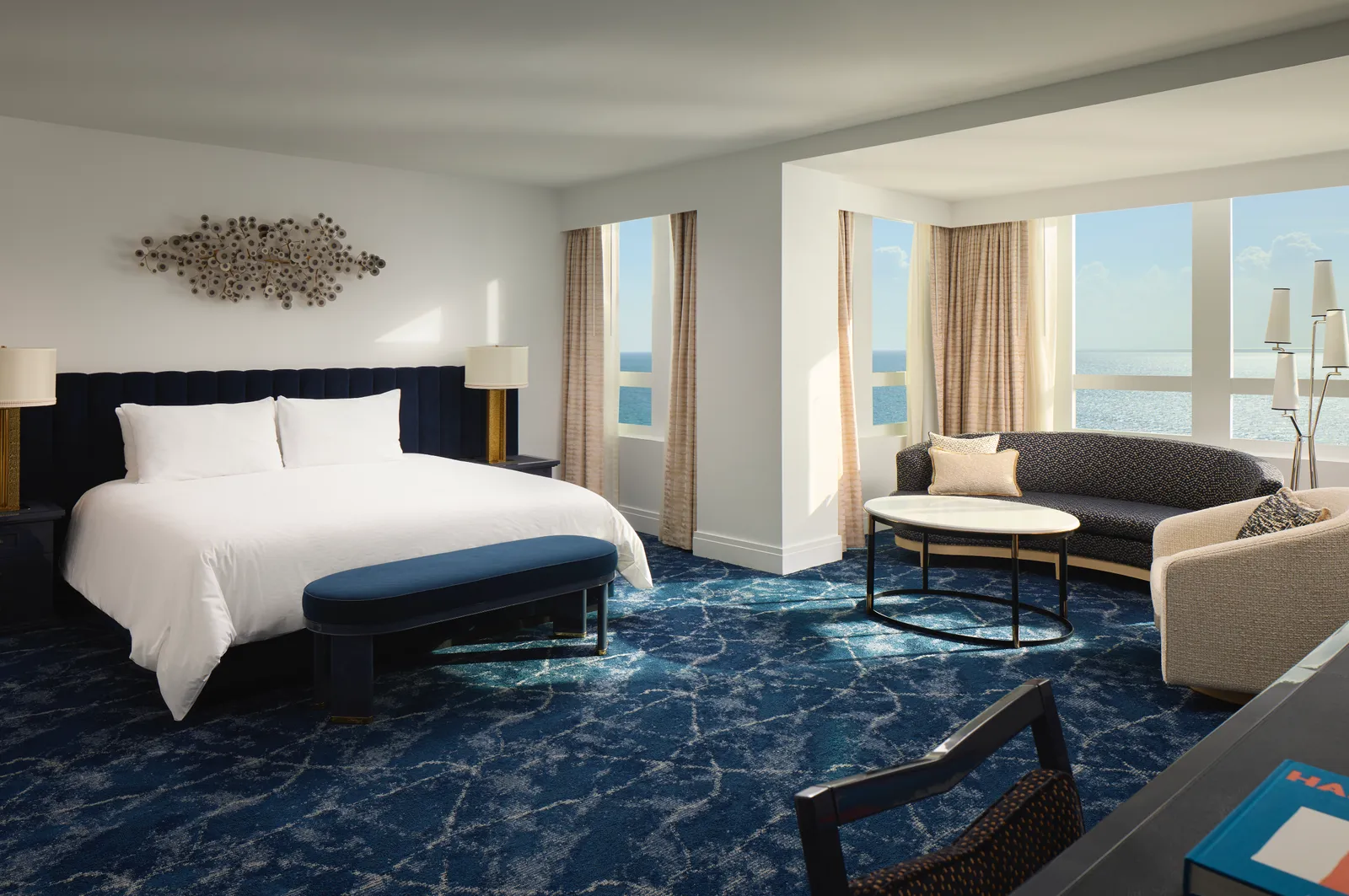 A bedroom of the Bleau Royale Suite, with the bed, a blue bench at its foot, a sofa and the windows over the ocean.