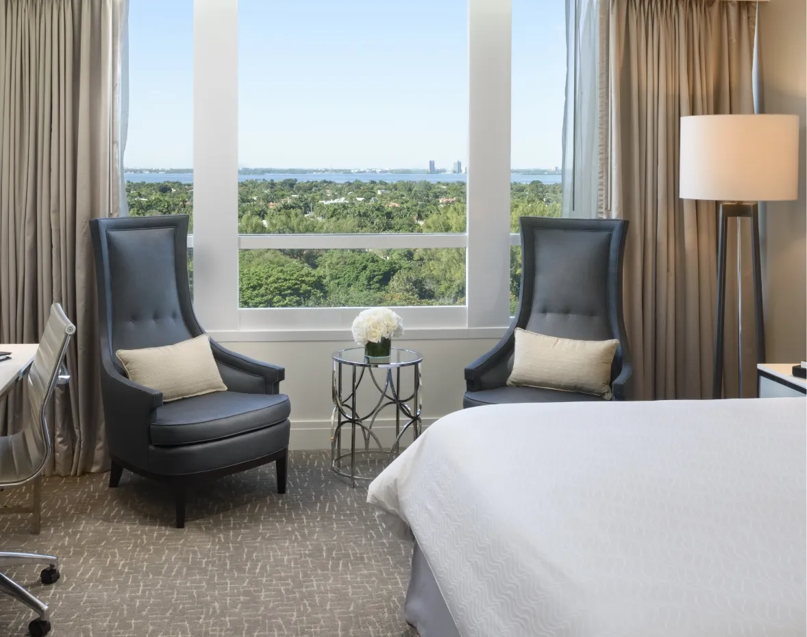 A Deluxe Bay View room: 2 blue armchairs and a side table by the window, which looks over trees to the bay, and the corner of the bed.