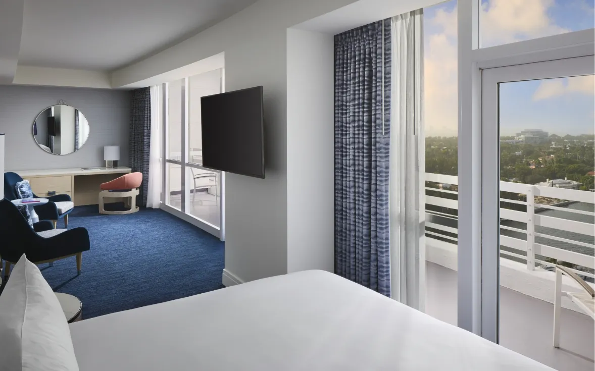 A Deluxe room with balcony: the bed, blue carpet, the desk with a round mirror and the door open onto the balcony and the trees.