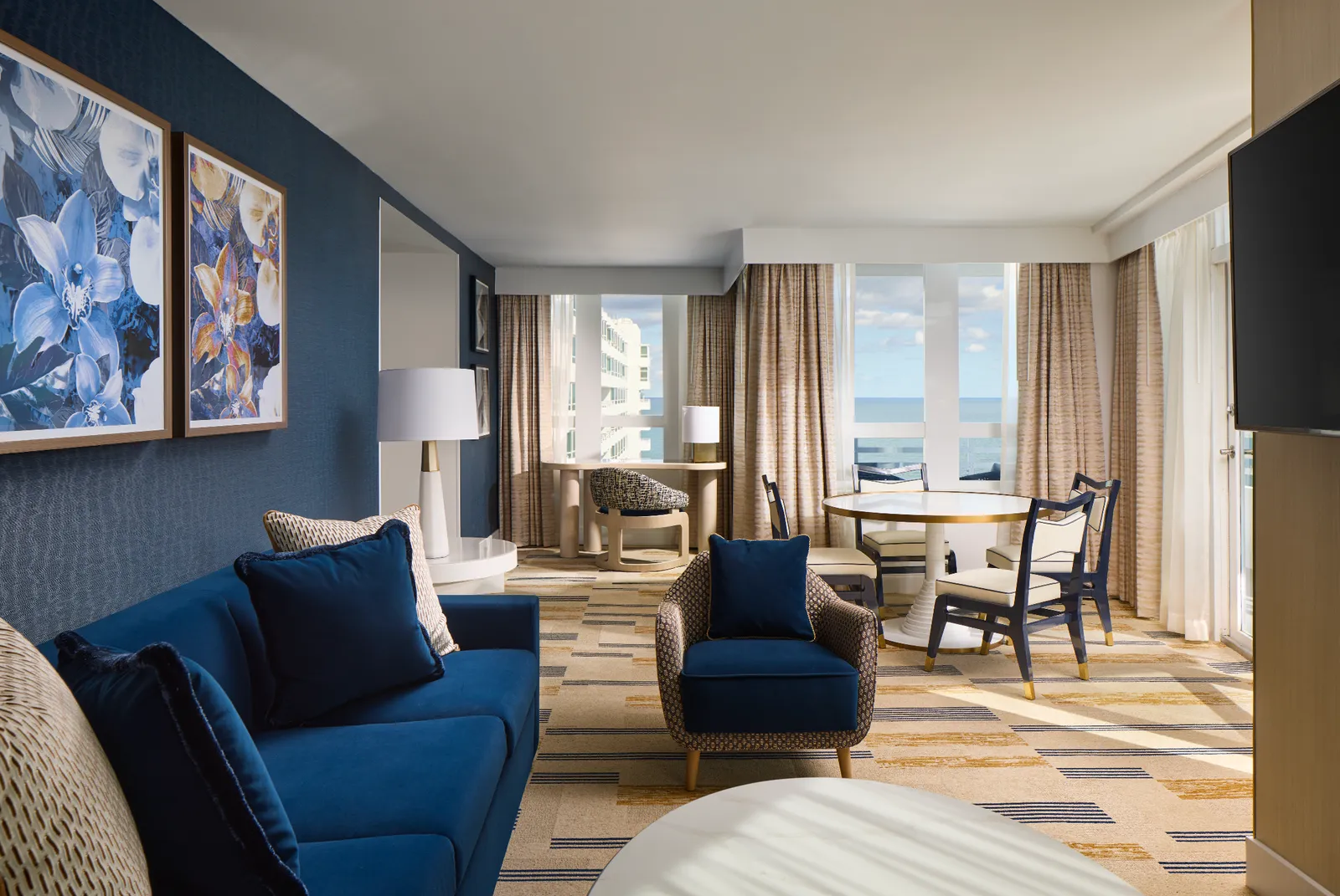 The Executive One Bedroom Suite’s living room: blue sofa and armchairs, 2 floral paintings, the round dining table and corner windows.