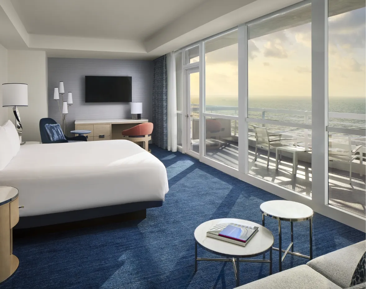 An Oceanfront Junior Suite: the bed, round side tables and the glass wall onto the balcony and the ocean in morning light.