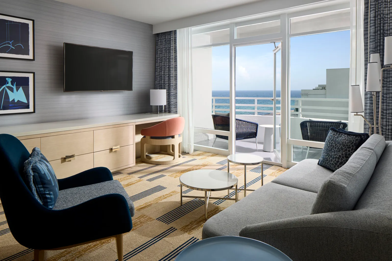 The living room of an Oceanfront One Bedroom Suite: a grey sofa, a blue armchair, the desk and the door to the balcony and the ocean.