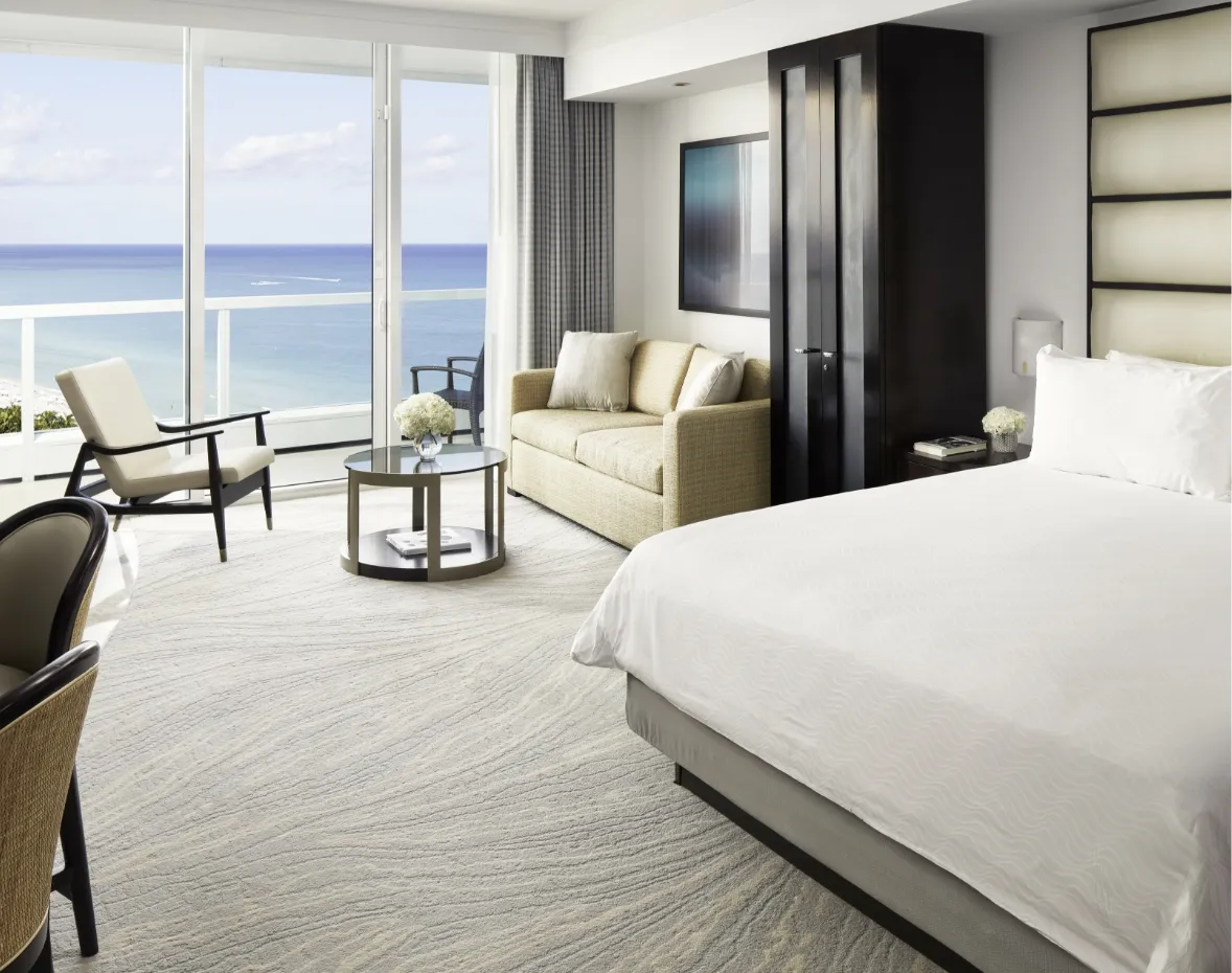 A Sorrento tower Junior Suite: the bed, a pale sofa with 2 armchairs and the glass doors onto the balcony and the sea.