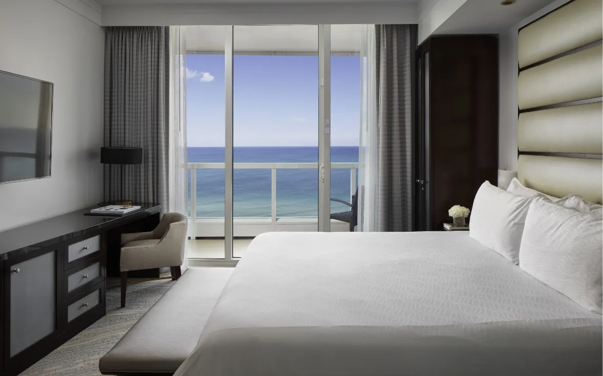 The bedroom of a Sorrento Oceanfront One Bedroom Suite, with its king bed, the bench at its foot and the ocean beyond the glass.