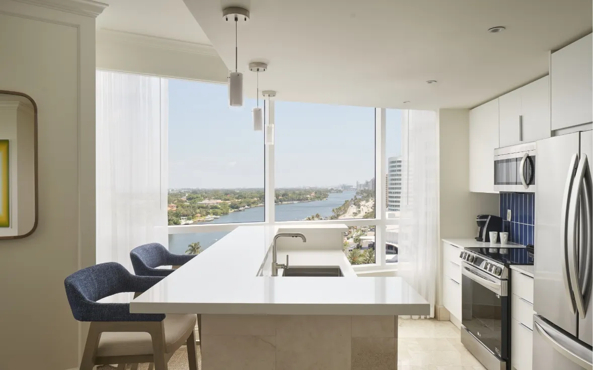 The kitchen of a Trésor Bay View One Bedroom Suite: the marble island with stools, steel appliances and the view along the waterway and the coast.