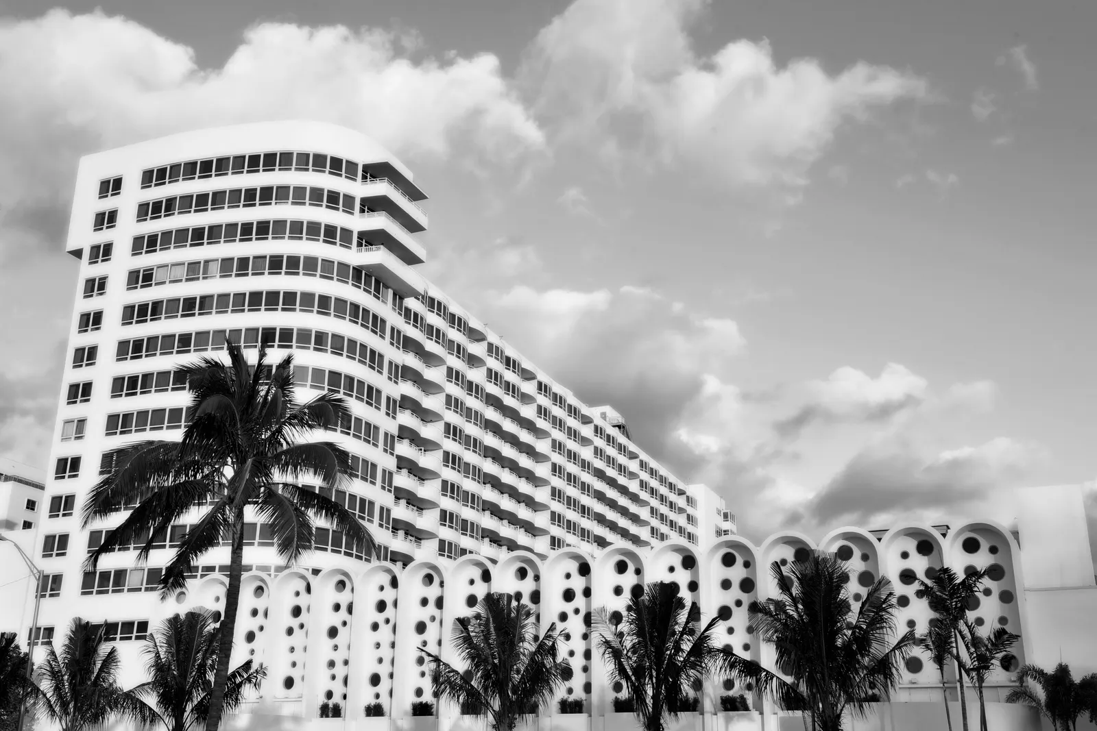 In black and white, the hotel’s long curved façade with its rows of balconies, palms and, at its base, the arched wall pierced by round holes.