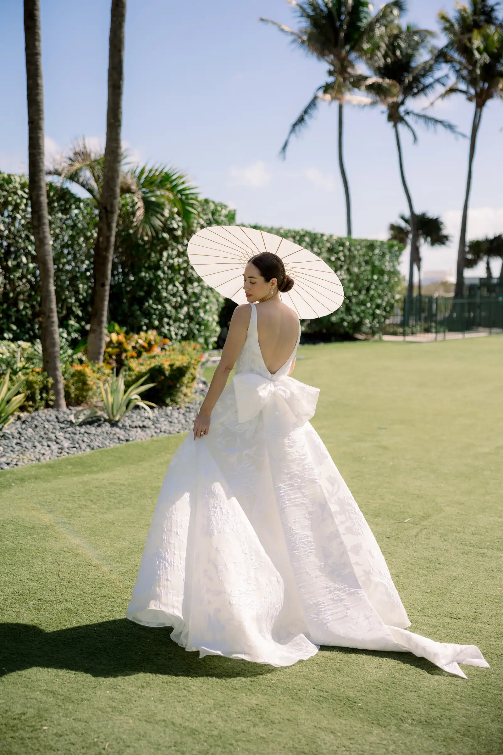 A bride seen from behind with a white parasol on a lawn, among palms and hedges.