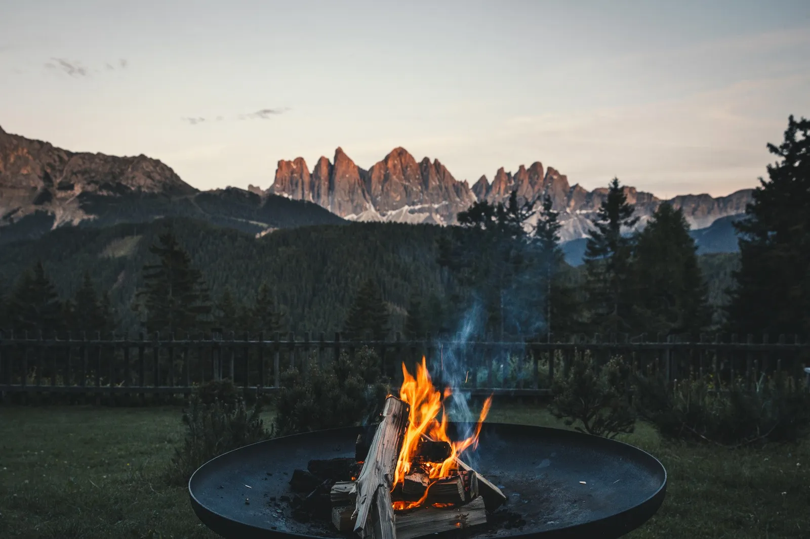A fire bowl burning on the lawn at dusk, with the fence, the firs and the pink spires of the Odle behind.