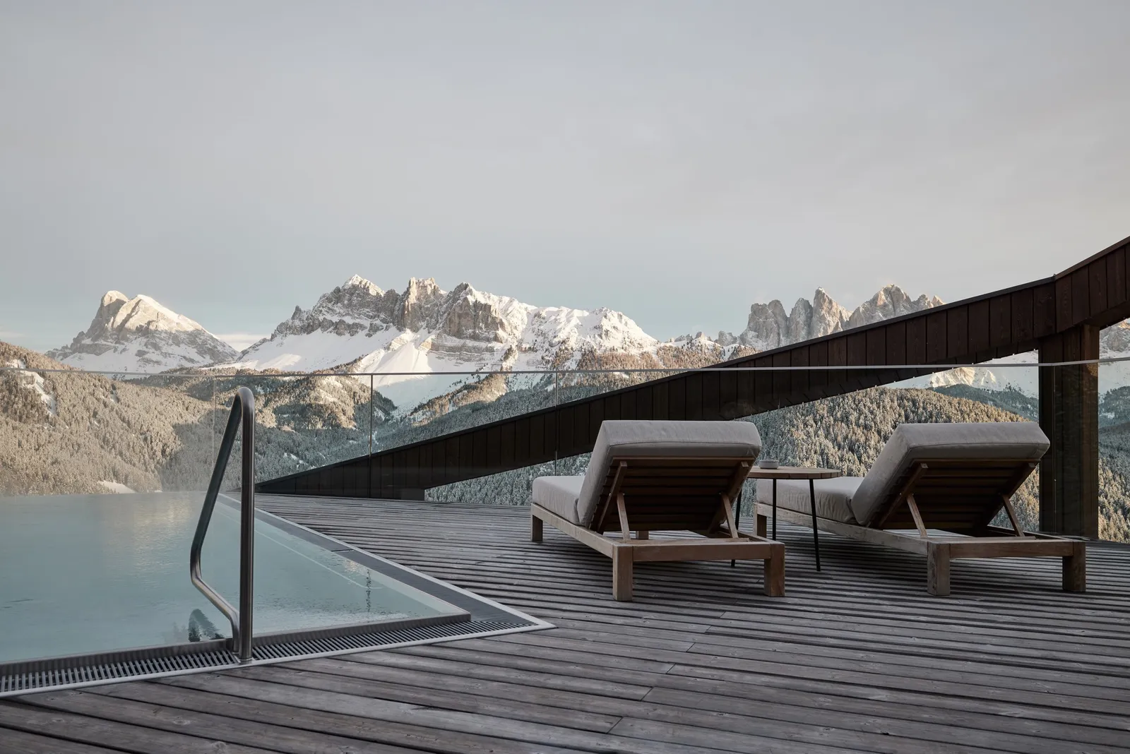 A Penthouse rooftop pool, with 2 loungers on the timber deck and the snow-covered Dolomites beyond the glass balustrade.