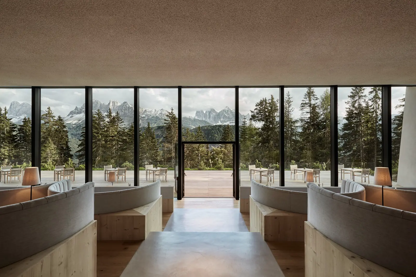 The terraced restaurant: curved wood-and-fabric booths step down to the glass wall, with the terrace, the firs and the Dolomites beyond.