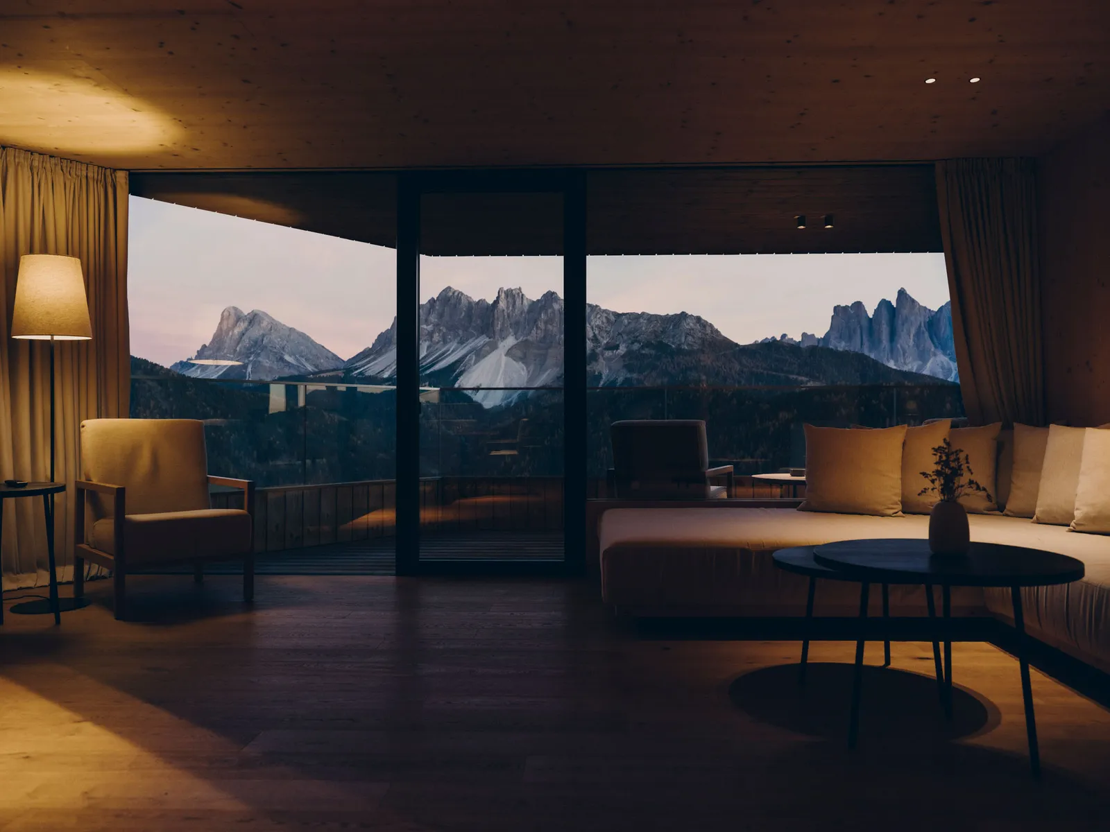 A Penthouse living room in the evening: the spruce ceiling, the sofa, the armchairs and the glass wall onto the terrace before the Dolomites.