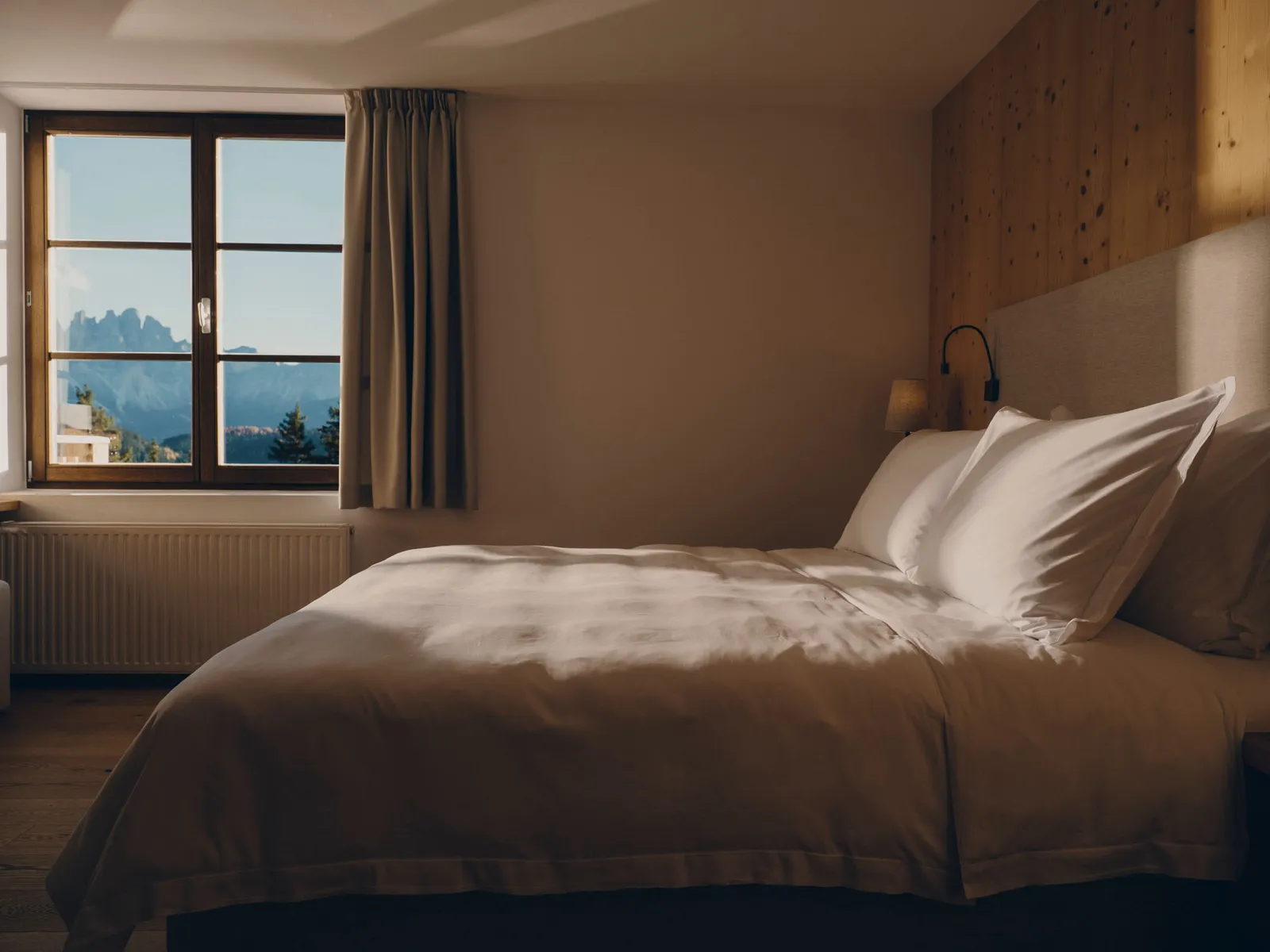 A Room: the bed with white linen, the wooden headboard and wall, and the window framing the Odle.