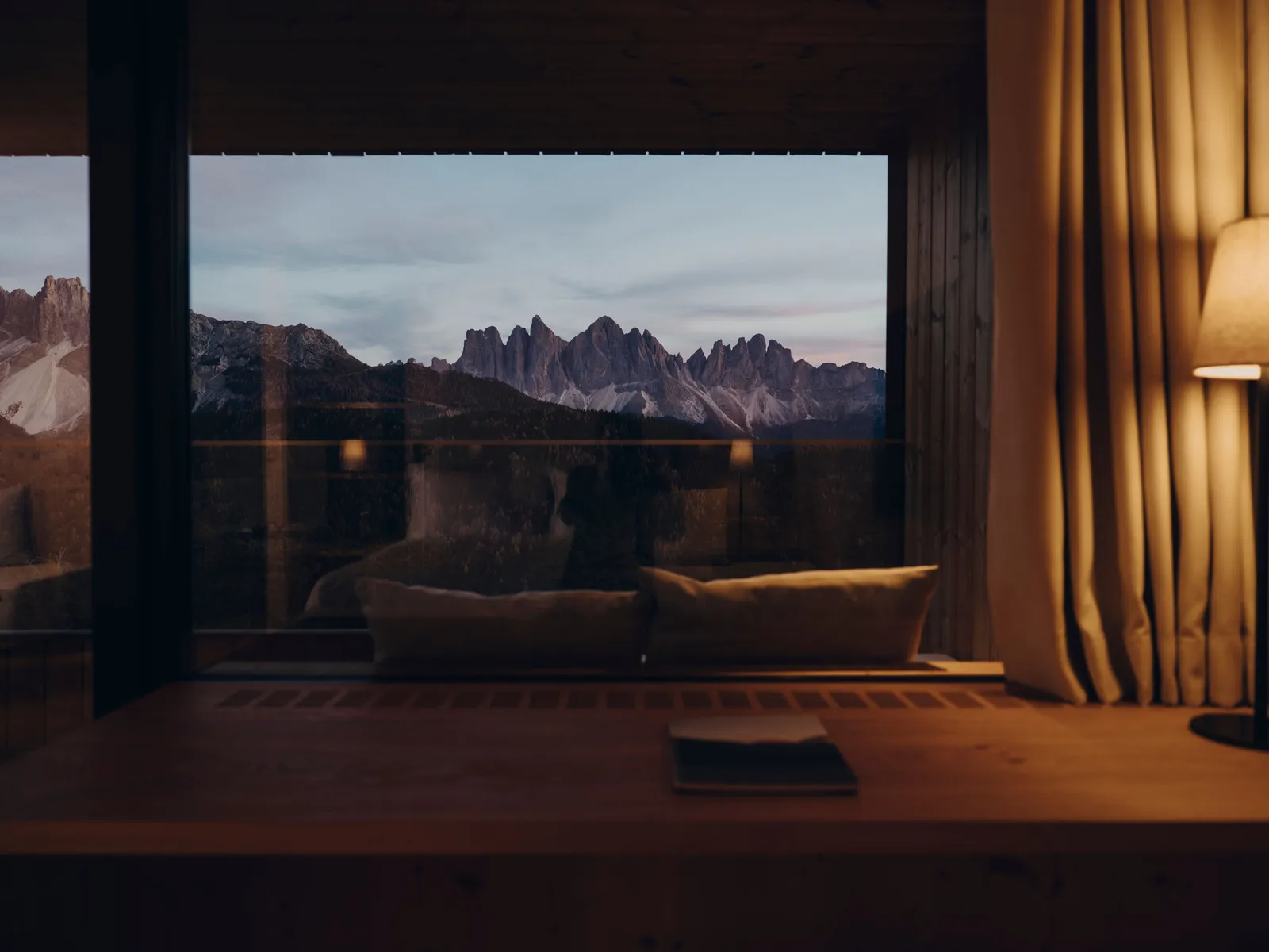 A Tower Suite at dusk: the wall-wide window onto the Odle, the daybed on the terrace and a lamp lit beside the curtain.