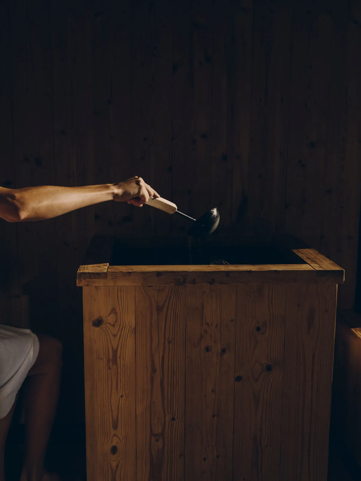 An arm ladles water onto the stones of a wood-clad stove in the half-light of the sauna.