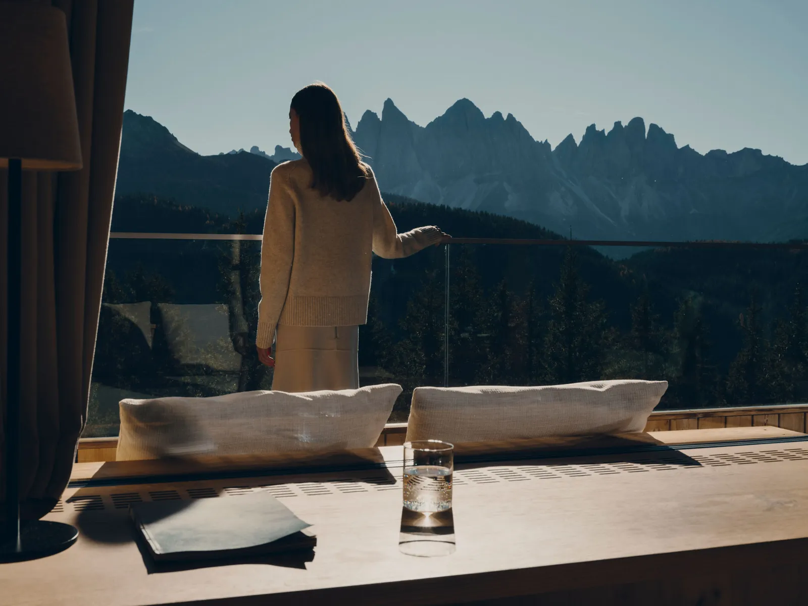 A woman seen from behind on a suite terrace, a hand on the glass balustrade, looks at the spires of the Odle; a daybed with 2 cushions in front.