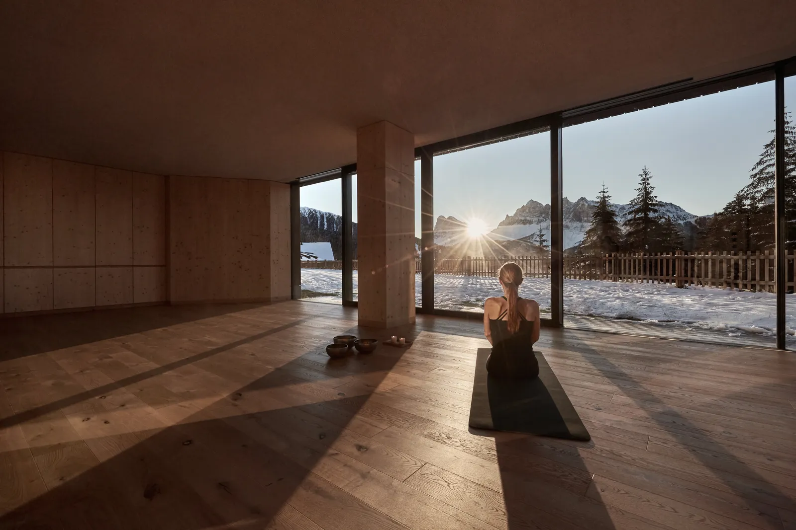 The Wyda room: a woman seated on a mat before the glass wall, the low sun above the snowy mountains.