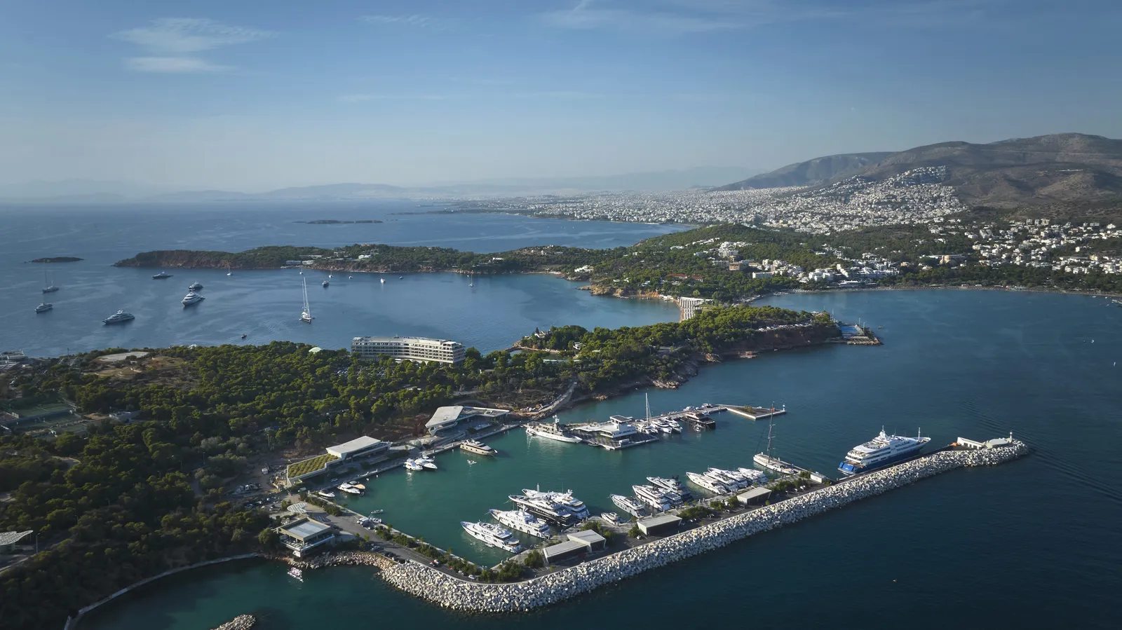 Astir Marina from the air, with yachts moored behind the breakwater, the pine-covered peninsula and the coast towards Athens.