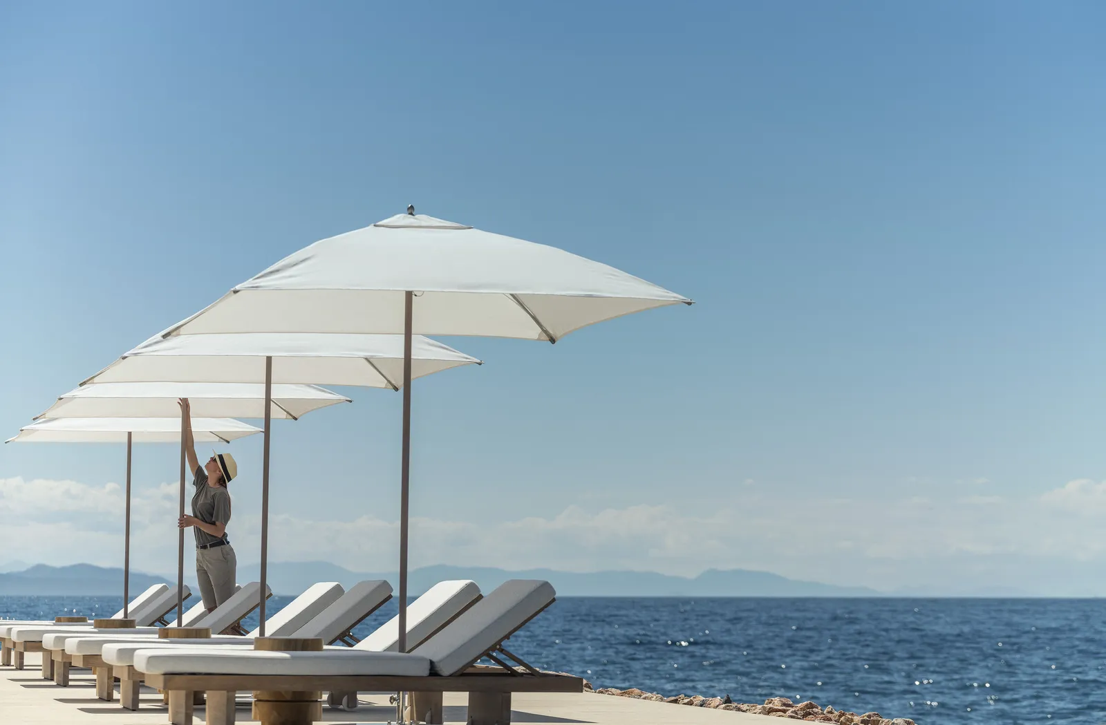 A row of loungers under large white umbrellas at the water’s edge, an attendant opening one of them; islands on the horizon.