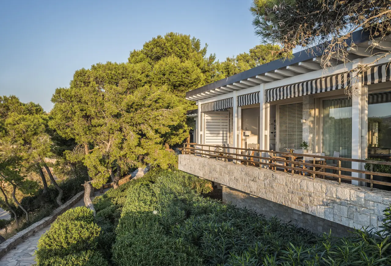 A bungalow on a stone base among the pines, with blue-and-white striped awnings and the paved path running down towards the sea.