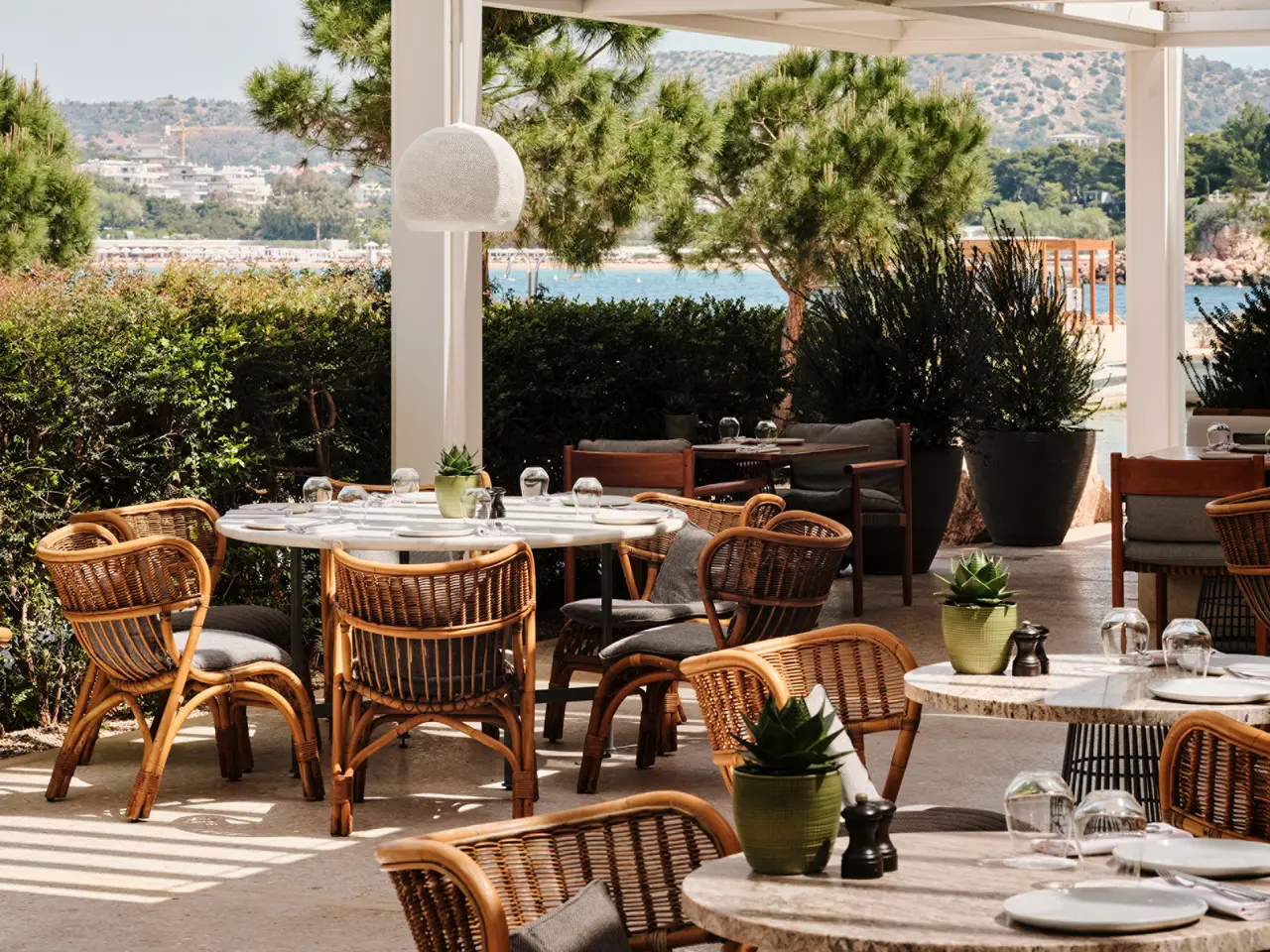 The terrace of Helios, with travertine tables, rattan chairs, potted herbs and the bay beyond the hedge.