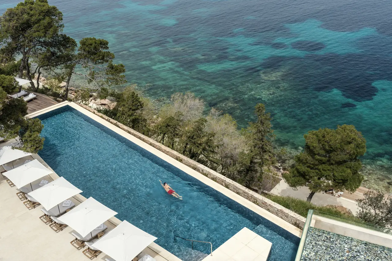 The Arion pool from above, long and narrow over the sea, with one swimmer and a row of loungers under white umbrellas.