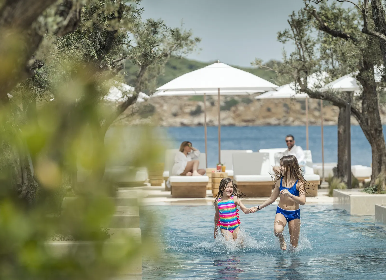 2 girls run hand in hand through the shallows of the Nafsika pool, among olive trees, with umbrellas and the bay behind.