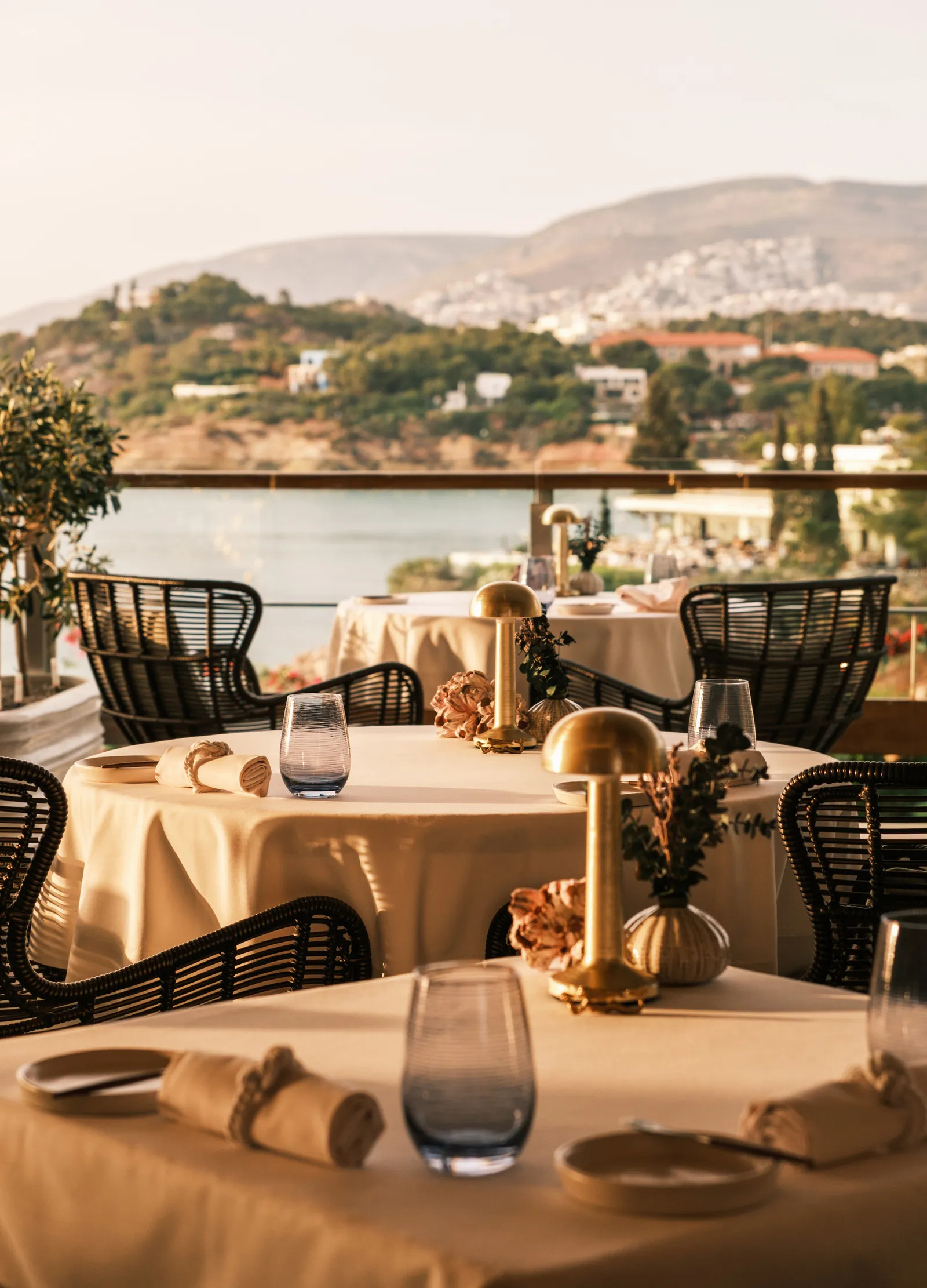 A laid table on the terrace of Pelagos at sunset, with pale linen, brass lamps and the hills of Vouliagmeni across the bay.