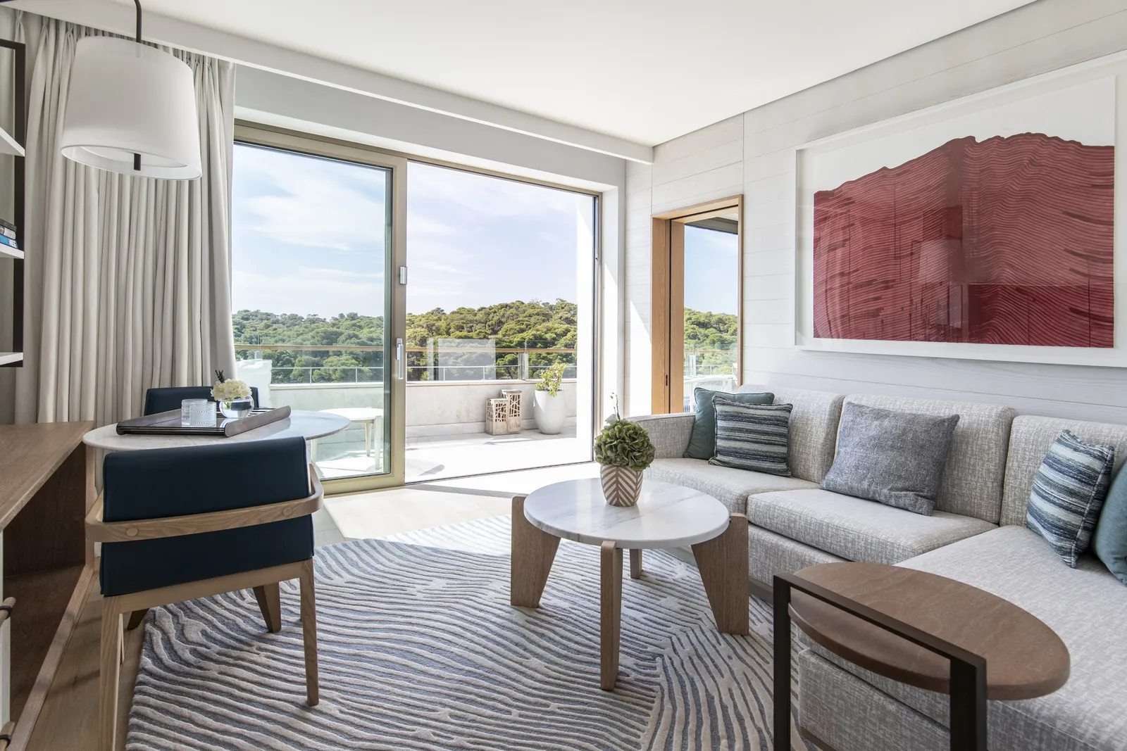 The sitting room of an Arion Executive Suite, with a corner sofa under a red painting and glass doors open to the balcony and the pines.