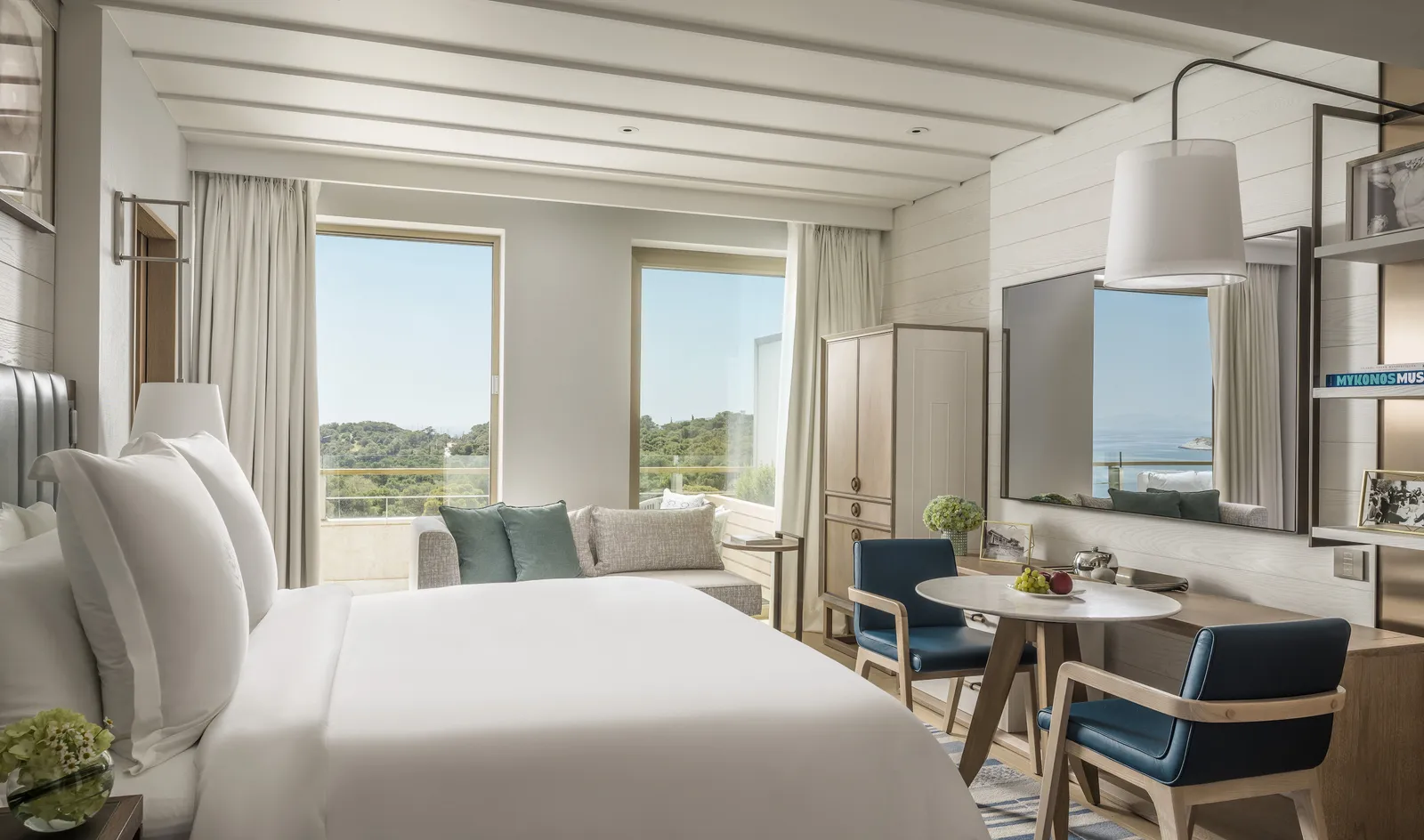 An Arion room with a white bed, a sofa under 2 windows onto the pines and a round table with blue chairs.