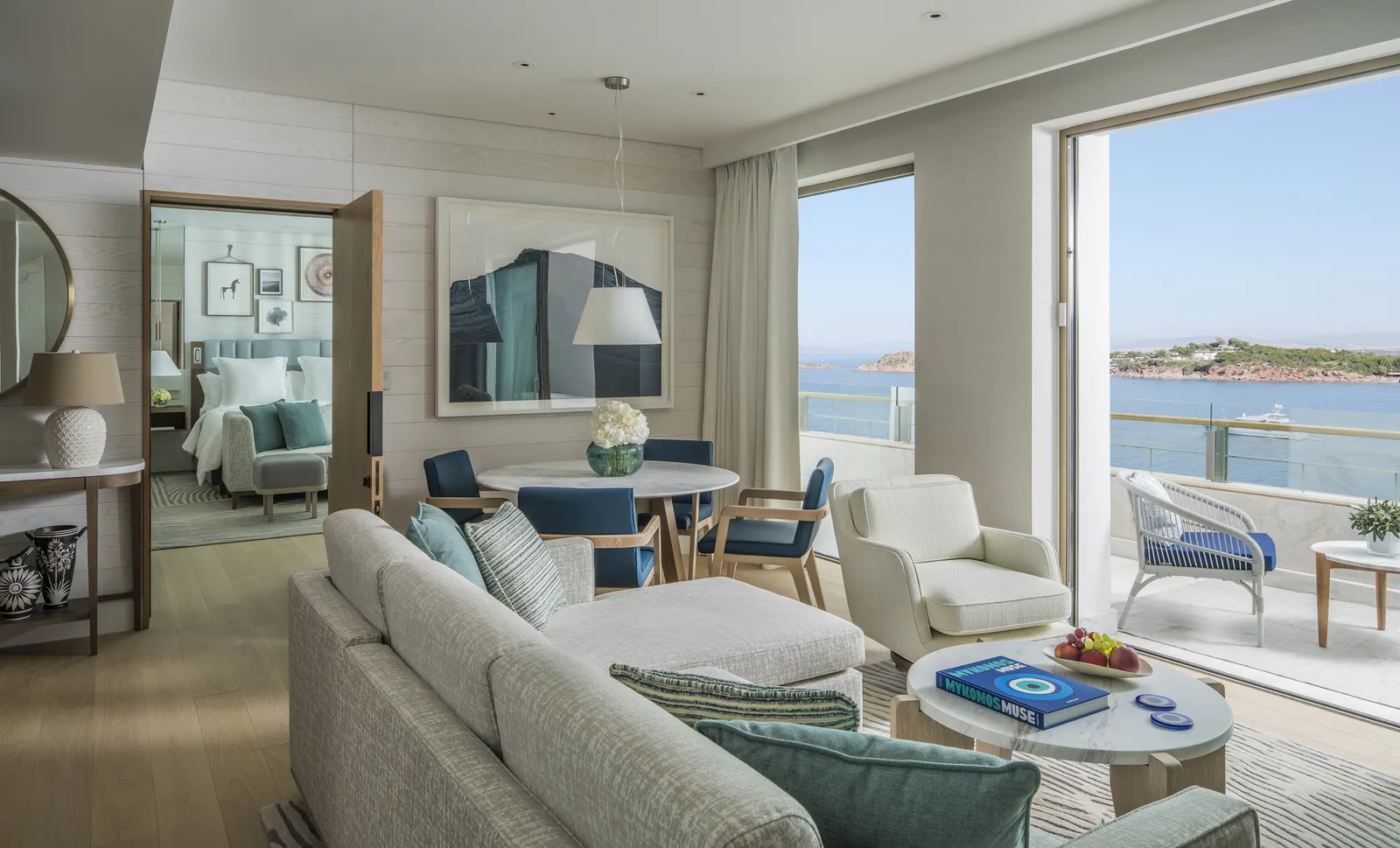 The living room of an Arion one-bedroom suite: pale sofas, a dining table, the bedroom through the door and the balcony over the sea.