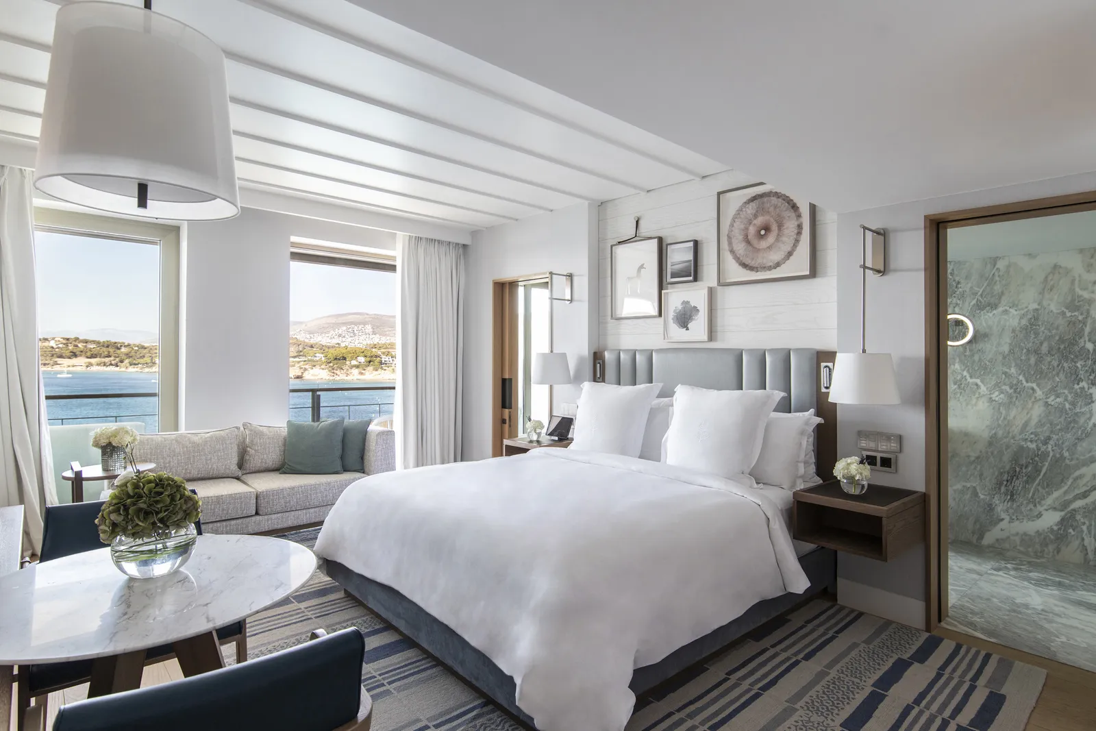 An Arion sea-view room: the bed with its grey headboard, a sofa along the windows onto the bay and the glass door of the marble bathroom.