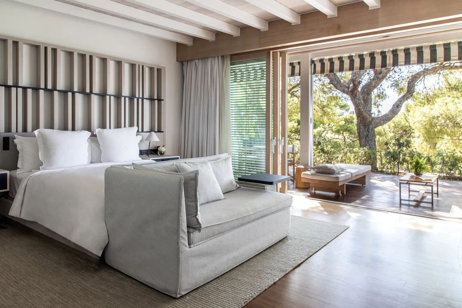 A garden-view bungalow: the bed and a sofa facing open glass doors, a striped awning and the timber terrace under the pines.