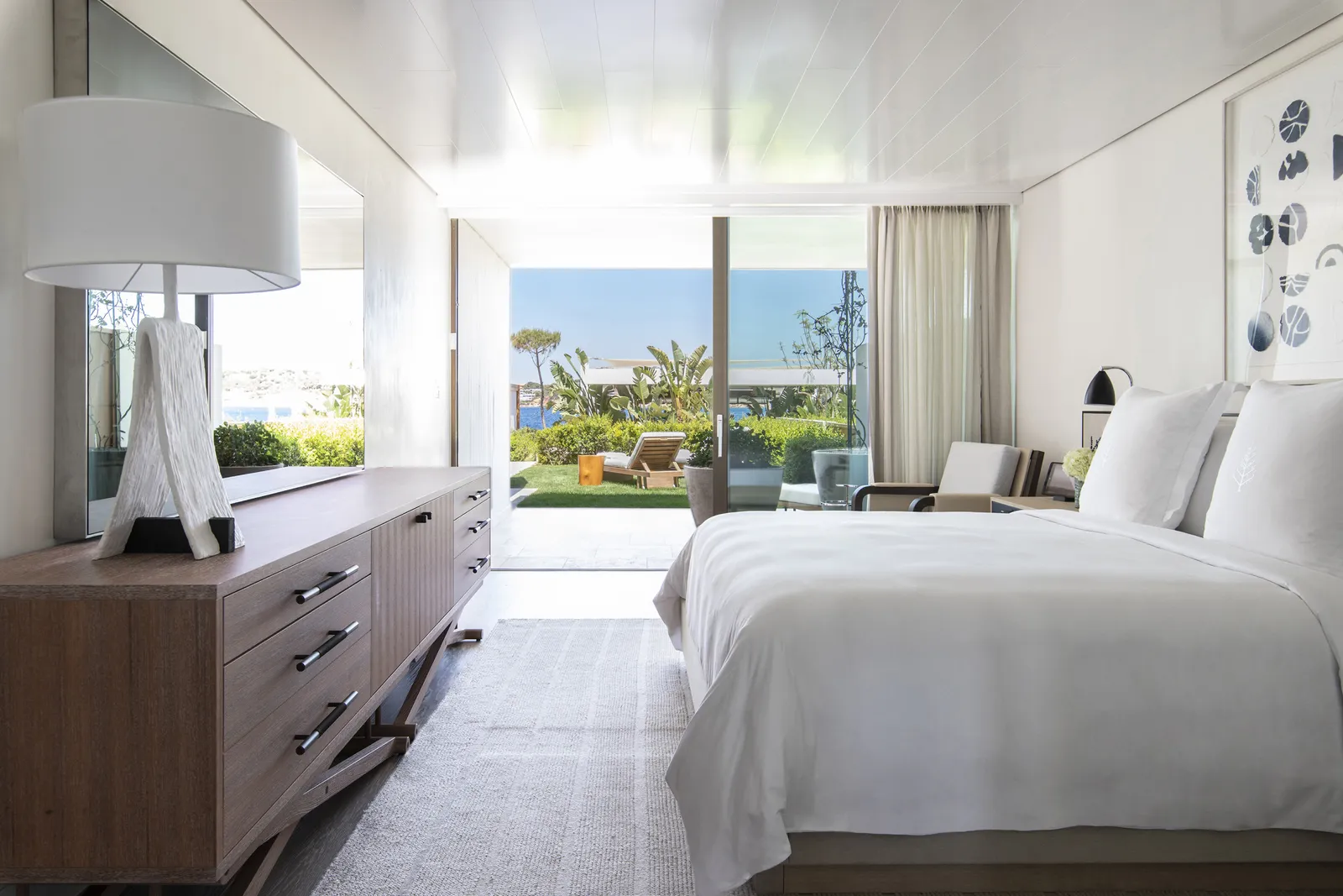 A ground-floor Nafsika room: the bed, a timber chest of drawers and the glass wall open to the garden with its loungers and the sea.
