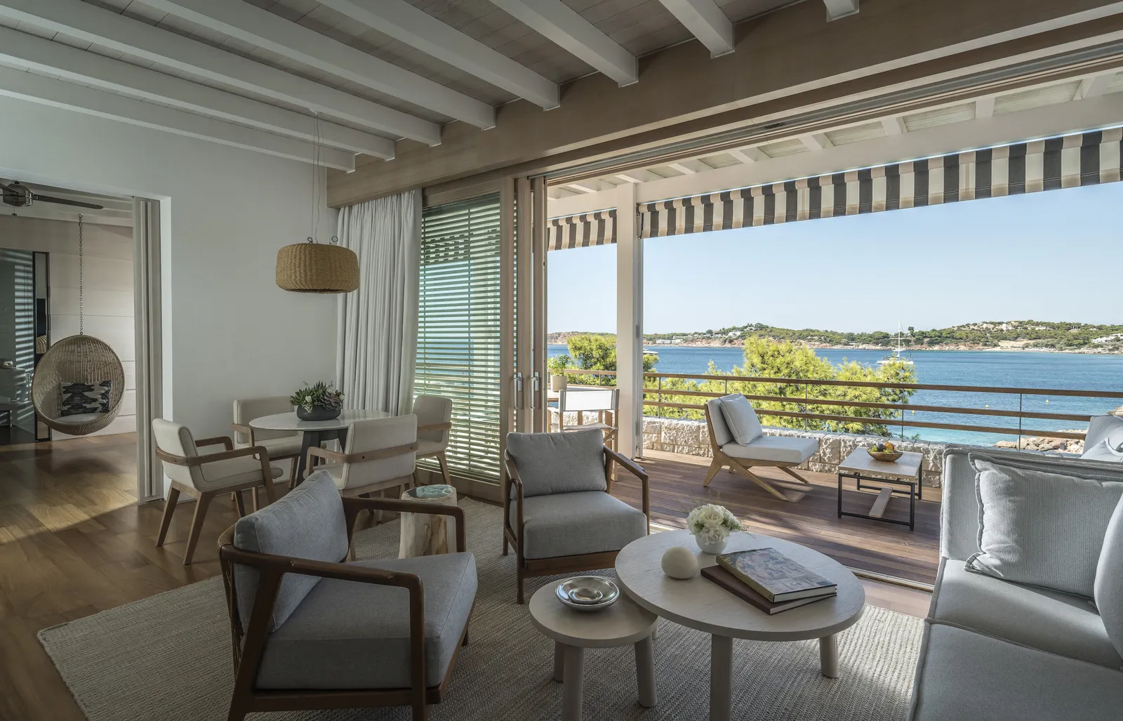 The living room of a Bungalow Suite, with armchairs, a dining table and glass doors open to the covered terrace and the sea.