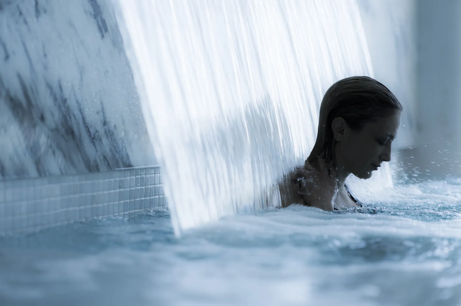 A woman under the waterfall jet of the spa’s hydrotherapy pool, between walls of pale marble.
